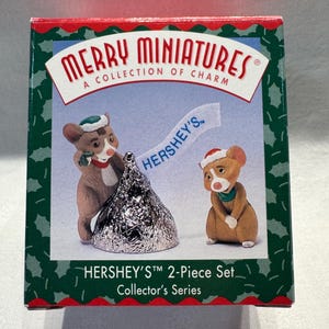 May include: A Hershey's Merry Miniatures 2-piece collector's set box featuring two brown mice wearing red and green holiday hats and scarves. One mouse stands beside a silver Hershey's Kiss, while the other stands nearby. The box has a holly border.