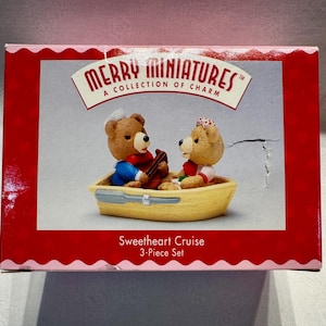 May include: A Merry Miniatures Sweetheart Cruise 3-Piece Set box featuring two brown teddy bears in a yellow boat. One bear wears a blue jacket and white hat, while the other wears a red and white patterned headpiece. The box has red borders.