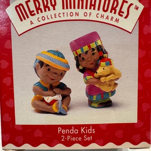 May include: A Merry Miniatures Penda Kids 2-Piece Set box featuring two small figurines. One figurine sits playing a drum, while the other stands holding a yellow bear. The packaging is red with white heart patterns and includes the text Merry Miniatures.