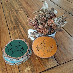 May include: Wooden coasters with painted designs. One is green with black sprigs, the other orange with black leaf designs. A small white mug with dried flowers is in the background.