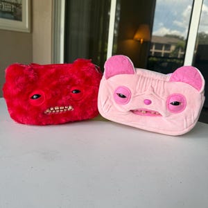 May include: Two rectangular plush pouches, one bright red and one light pink. Each pouch features a creature-like face with prominent, realistic-looking teeth, heavy-lidded eyes, and small ears. The red pouch has a fuzzy texture, while the pink pouch has a smooth, soft fabric finish with stitched details around the eyes and nose.