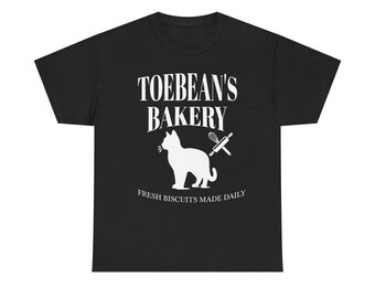 May include: Black t-shirt with a white graphic featuring a cat silhouette, rolling pin, and the text "TOEBEAN'S BAKERY." Below the bakery name, the text reads "FRESH BISCUITS MADE DAILY." The design is centered on the shirt.