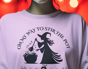 Könnte beinhalten: Ein helllila Sweatshirt mit dem Text "ON MY WAY TO STIR THE POT", der eine schwarze Grafik einer Hexe umkreist, die einen Kessel umrührt. Das Design beinhaltet Fledermäuse und einen Totenkopf mit einem Kessel.