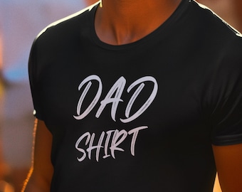 Peut inclure: T-shirt noir avec l'inscription blanche "DAD SHIRT" dans un style manuscrit. Le t-shirt est à col rond et semble être fait d'une matière douce. L'image est un gros plan du t-shirt.