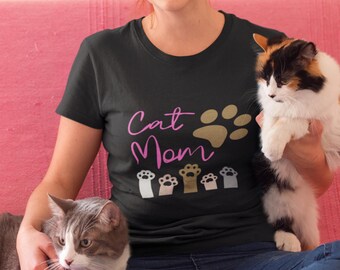 Peut inclure: T-shirt noir avec l'inscription "Cat Mom" en rose et des empreintes de pattes dorées. Le t-shirt présente divers motifs d'empreintes de pattes en blanc, beige et marron. Un chat tricolore et un chat gris et blanc sont également visibles.