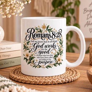 May include: A white ceramic mug featuring a decorative floral wreath design in green and gold. The text reads: Romans 8:28 And we know that in all things God works for the good of those who love him, who have been called according to his purpose.