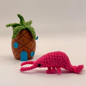 Può includere: Una casa marrone a forma di ananas con una parte superiore verde all'uncinetto e dettagli blu, accanto a un gamberetto rosa all'uncinetto. La casa ha una porta e finestre. Il gamberetto ha occhi neri e un corpo curvo.