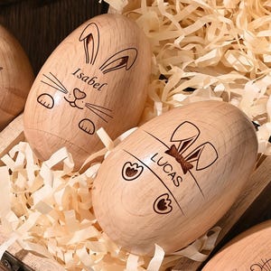 May include: Wooden Easter eggs with engraved bunny designs and names, presented in a wooden box with shredded paper. The eggs feature names such as "Isabel" and "Lucas", and the box is on a wooden surface.