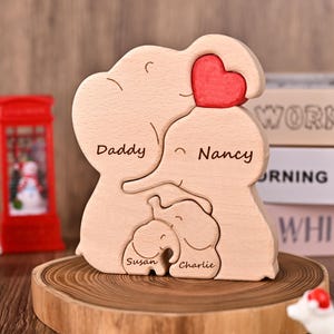 Può includere: Un puzzle in legno a forma di famiglia di elefanti con i nomi "Daddy", "Nancy", "Susan" e "Charlie" incisi. Il puzzle presenta un cuore rosso ed è esposto su una fetta di legno. La famiglia di elefanti è realizzata in legno chiaro.