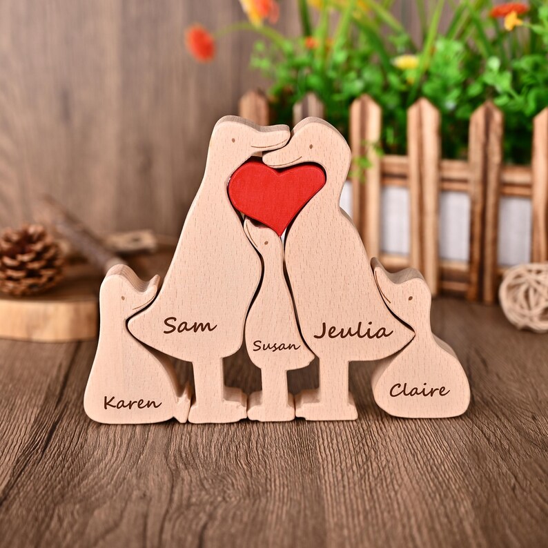 May include: A wooden family sculpture of five duck-shaped figures. The central ducks form a heart shape with a red heart insert. Names are engraved on each duck: Karen, Sam, Susan, Jeulia, and Claire. The sculpture is made of light-coloured wood.