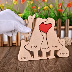 May include: A wooden family puzzle in the shape of a duck family. The puzzle has a red heart and is engraved with the names "Dad", "Mom", "Susan", "Jeulia", and "Karen". The puzzle is made of light-coloured wood.