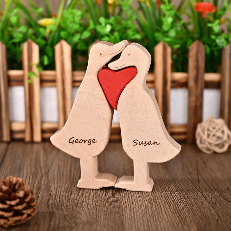 May include: Wooden duck figurine with a red heart. The ducks are light brown with the names "George" and "Susan" engraved on them. The figurine is on a wooden surface with a small pine cone and a decorative ball.