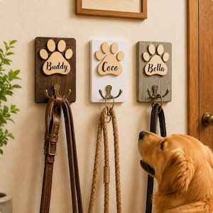 Peut inclure: Trois patères murales rectangulaires en bois avec un motif de patte gravé et les noms Buddy, Coco et Bella inscrits. Chaque support comporte une fixation métallique retenant une laisse en cuir, une laisse en corde et une laisse noire.