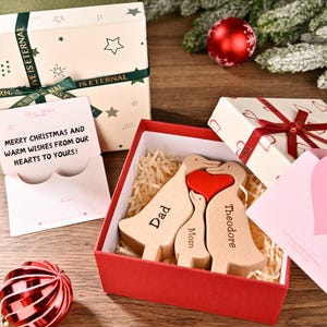 May include: A wooden duck family with a red heart, presented in a red box. The ducks are engraved with "Dad", "Mom", and "Theodore". A gift box and a card with the message "Merry Christmas and warm wishes from our hearts to yours!" are also visible.