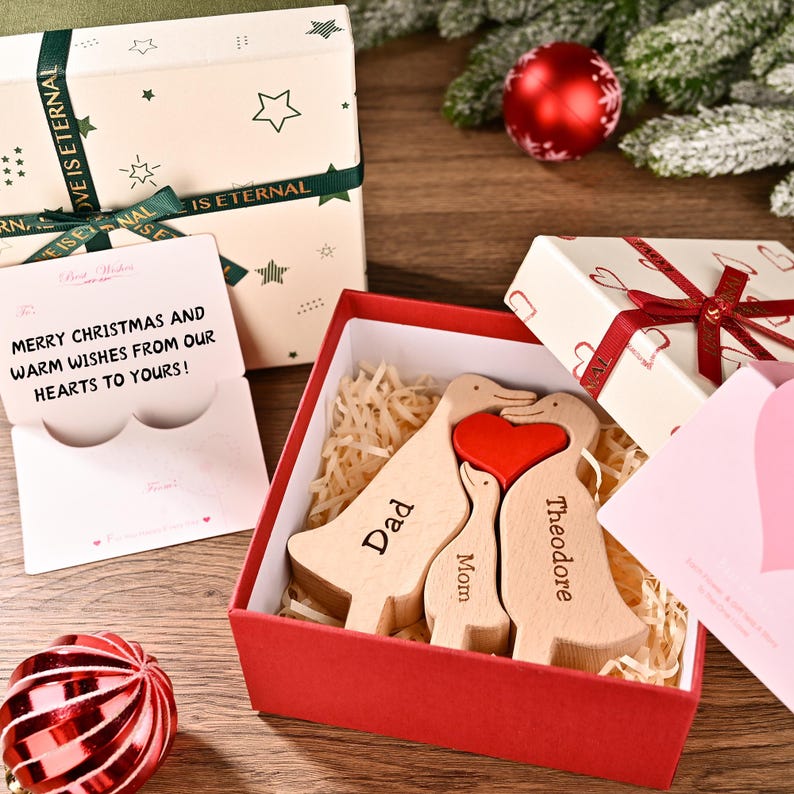 May include: A wooden duck family with a red heart, presented in a red box. The ducks are engraved with "Dad", "Mom", and "Theodore". A gift box and a card with the message "Merry Christmas and warm wishes from our hearts to yours!" are also visible.