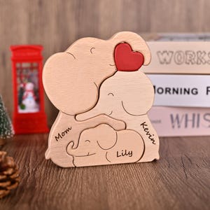 Puede incluir: Un rompecabezas de madera con forma de tres elefantes, con un coraz&oacute;n rojo. El rompecabezas est&aacute; grabado con los nombres "Mom", "Kevin" y "Lily". Los elefantes son de color marr&oacute;n claro.