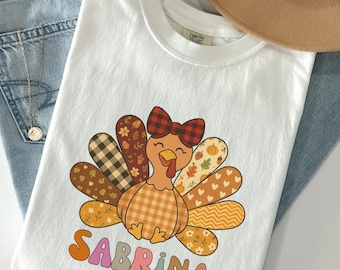 Könnte beinhalten: Weißes T-Shirt mit einem Cartoon-Truthahn-Design, einer karierten Schleife und gemusterten Schwanzfedern. Der Name "SABRINA" ist in einer Retro-Schriftart aufgedruckt. Das Shirt liegt auf einer blauen Jeans und einem beigen Hut.