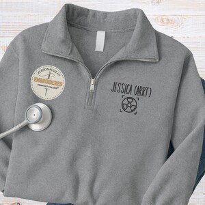 May include: A gray Comfort Colors quarter-zip sweatshirt with the name "JESSICA (ARRT)" and a radiation symbol printed on the front. The sweatshirt has a personalized embroidered patch and a stethoscope.