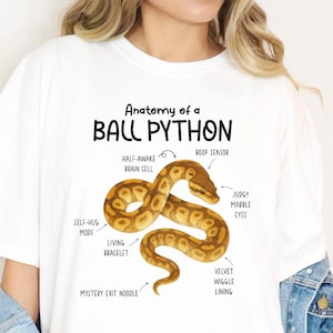 May include: White t-shirt featuring a detailed illustration of a ball python with labeled anatomical features. The text "Anatomy of a BALL PYTHON" is printed above the snake graphic. The snake is brown and yellow.
