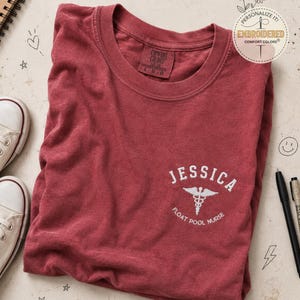 May include: A dark red short-sleeve t-shirt with a crew neckline. The chest features white embroidery reading JESSICA above a caduceus symbol and the text FLOAT POOL NURSE. The inner neck label displays the Comfort Colors brand and size L.