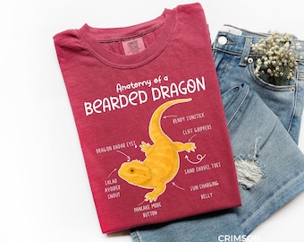 Op de afbeelding: Karmizijnrood T-shirt met een gele illustratie van een baardagaam en gelabelde lichaamsdelen. De tekst "Anatomy of a Bearded Dragon" staat erboven. Het T-shirt ligt naast een blauwe jeans.