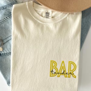 May include: Ivory-colored t-shirt with the word "BAR tender" embroidered in yellow. The shirt is folded and placed on a light-colored surface, with a tan hat and denim jeans in the background.