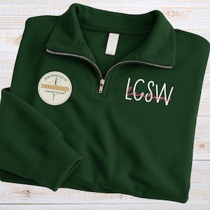 May include: A dark green quarter-zip sweatshirt with the embroidered text "LCSW" in white and pink. The sweatshirt has a white tag and a circular patch that says "Personalize it! Embroidered Comfort Colors". The zipper is silver.