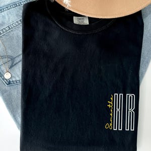 Peut inclure: T-shirt noir plié avec le texte "Samantha HR" en design vertical. Le mot "Samantha" est en écriture jaune et "HR" en lettres blanches. Le t-shirt est posé sur un fond clair.