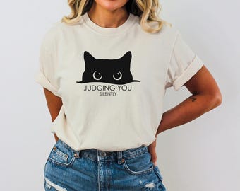 May include: Cream-colored t-shirt featuring a black cat graphic with the text "JUDGING YOU SILENTLY". The cat's eyes are visible above the text. The shirt is paired with blue denim jeans.