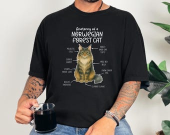May include: Black t-shirt featuring a detailed illustration of a Norwegian Forest Cat with labeled anatomical features. The text "Anatomy of a Norwegian Forest Cat" is printed above the cat image. The shirt is worn by a person.