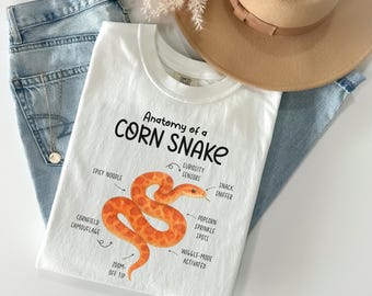 Puede incluir: Camiseta blanca con el texto "Anatomy of a Corn Snake" y una ilustración de una serpiente naranja. La camiseta incluye etiquetas como "Spicy Noodle" y "Popcorn Sprinkle Spots". La camiseta está colocada sobre unos vaqueros azules y junto a un sombrero beige.
