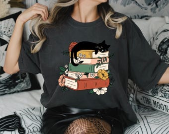 Könnte beinhalten: Dunkelgraues T-Shirt mit einer Grafik einer schwarzen Katze, die auf einem Stapel bunter Bücher schläft. Die Bücher sind mit floralen Akzenten in Rot, Gelb und Weiß verziert. Ein lässiges, bequemes T-Shirt.
