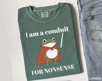 May include: A green t-shirt with the text "I am a conduit for nonsense." The shirt features a cartoon frog wearing a red cape and holding a sword. The shirt is next to a pair of blue jeans.
