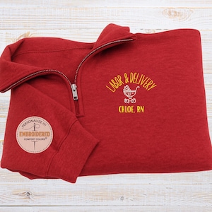 Könnte beinhalten: Ein rotes Sweatshirt mit Viertelreißverschluss und dem gestickten Text "LABOR & DELIVERY" sowie einer gelben Babywagen-Grafik. Der Text "CHLOE, RN" steht darunter. Das Sweatshirt hat auch einen kleinen gestickten Aufnäher mit der Aufschrift "PERSONALIZE IT! EMBROIDERED COMFORT COLORS®".