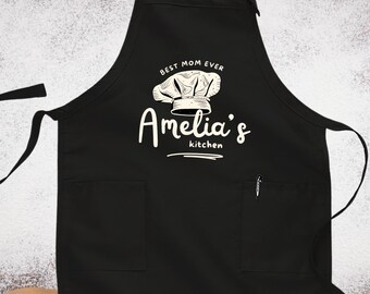 Op de afbeelding: Zwarte schort met witte tekst en een koksmutsymbool. De tekst luidt "Best Mom Ever" boven "Amelia's kitchen". De schort heeft twee voorzakken en een pen. De achtergrond is een houten oppervlak met wit poeder.