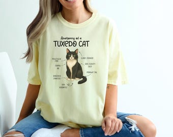 以下が含まれることがあります： タキシードキャットのグラフィックと解剖学的ラベルが描かれた、薄い黄色のTシャツ。「Anatomy of a Tuxedo Cat」の文字が猫のイラストの上にあります。猫は白と黒です。