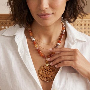 Puede incluir: Collar de varias hebras con un colgante grande y tallado. El collar incluye cadenas doradas y hebras de cuentas en tonos naranja, marrón y blanco. El colgante es de color marrón cálido con tallados detallados.