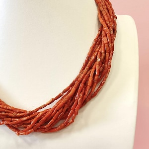Pode incluir: Um colar de v&aacute;rias voltas composto por pequenas contas cil&iacute;ndricas, vermelho-alaranjadas. O colar &eacute; exibido em um busto branco sobre um fundo rosa suave. As contas parecem estar enfiadas, criando um efeito em camadas.