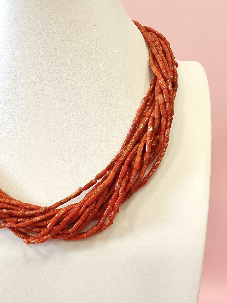 Pode incluir: Um colar de v&aacute;rias voltas composto por pequenas contas cil&iacute;ndricas, vermelho-alaranjadas. O colar &eacute; exibido em um busto branco sobre um fundo rosa suave. As contas parecem estar enfiadas, criando um efeito em camadas.