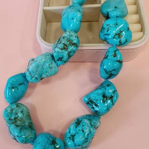 Puede incluir: Un collar compuesto por piedras de turquesa grandes, irregulares y pulidas que presentan vetas de matriz de color marr&oacute;n oscuro. Las piedras est&aacute;n ensartadas y aseguradas con un cierre de palanca de metal dorado, creando una pieza de joyer&iacute;a audaz.
