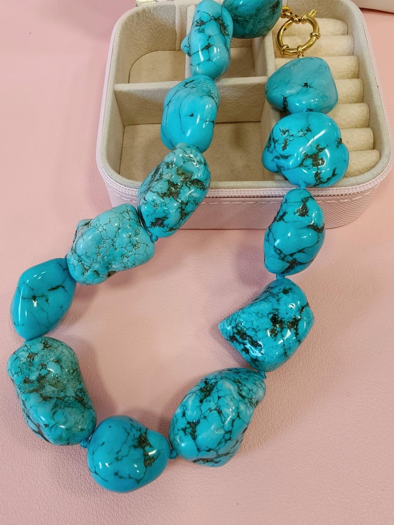 Puede incluir: Un collar compuesto por piedras de turquesa grandes, irregulares y pulidas que presentan vetas de matriz de color marr&oacute;n oscuro. Las piedras est&aacute;n ensartadas y aseguradas con un cierre de palanca de metal dorado, creando una pieza de joyer&iacute;a audaz.