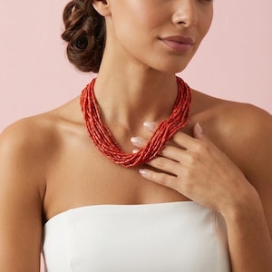 Pode incluir: Colar de coral vermelho de v&aacute;rias voltas. O colar &eacute; composto por pequenas contas cil&iacute;ndricas. A modelo est&aacute; usando um top branco sem al&ccedil;as. O fundo &eacute; de cor rosa lisa.