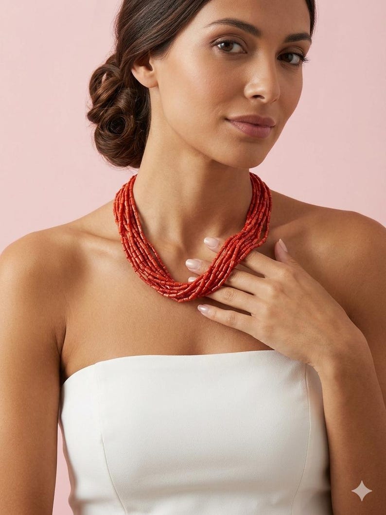 Pode incluir: Colar de coral vermelho de v&aacute;rias voltas. O colar &eacute; composto por pequenas contas cil&iacute;ndricas. A modelo est&aacute; usando um top branco sem al&ccedil;as. O fundo &eacute; de cor rosa lisa.