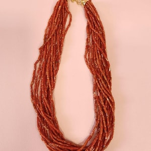 Pode incluir: Um colar de v&aacute;rias voltas composto por pequenas contas vermelho-alaranjadas. O colar &eacute; preso com um fecho dourado e tem uma apar&ecirc;ncia delicada e elegante. As contas s&atilde;o enfiadas juntas, criando um efeito de cascata.
