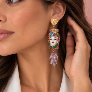 Può includere: Un orecchino color oro con una scultura di testa in ceramica decorata con una corona floreale, labbra rosse e un colletto verde. Il gioiello è completato da un perno a forma di foglia dorata e da un grappolo pendente di perline lilla traslucide.