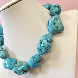 Puede incluir: Un collar que presenta grandes piedras de turquesa pulidas e irregulares con vetas de matriz de color marr&oacute;n oscuro. Las vibrantes piedras azul verdosas est&aacute;n ensartadas en un dise&ntilde;o grueso, mostrando variaciones naturales en forma y textura.