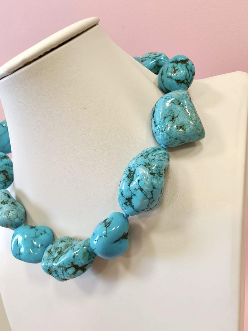 Puede incluir: Un collar que presenta grandes piedras de turquesa pulidas e irregulares con vetas de matriz de color marr&oacute;n oscuro. Las vibrantes piedras azul verdosas est&aacute;n ensartadas en un dise&ntilde;o grueso, mostrando variaciones naturales en forma y textura.