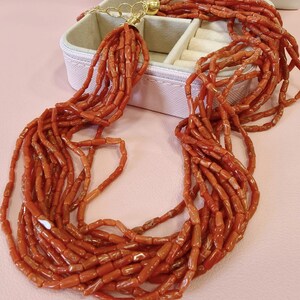 Pode incluir: Colar de v&aacute;rios fios com in&uacute;meros fios de contas de coral vermelho-alaranjado. O colar tem um fecho dourado e &eacute; exibido sobre uma superf&iacute;cie rosa ao lado de uma caixa de joias branca.