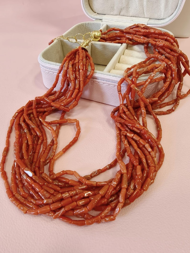 Pode incluir: Colar de v&aacute;rios fios com in&uacute;meros fios de contas de coral vermelho-alaranjado. O colar tem um fecho dourado e &eacute; exibido sobre uma superf&iacute;cie rosa ao lado de uma caixa de joias branca.