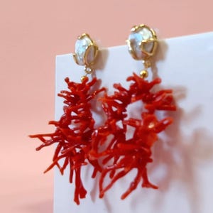Può includere: Un paio di orecchini ispirati al corallo. Ogni orecchino presenta un perno color oro con una perla bianca e un gruppo di elementi corallini rosso vivo a forma di ramo. Gli orecchini sono esposti su una superficie bianca con uno sfondo rosa.